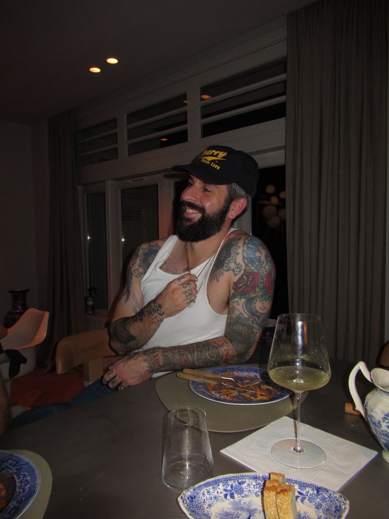 Joey de Boer laughing at a dinner table, tattooed arms, black and white